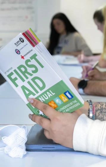 A close-up of a tattooed hand holding a first aid manual above a desk, with a white bandage and a tablet nearby—serious about learning, with a bit of style A close-up of a tattooed hand holding a first aid manual above a desk, with a white bandage and a tablet nearby—serious about learning, with a bit of style